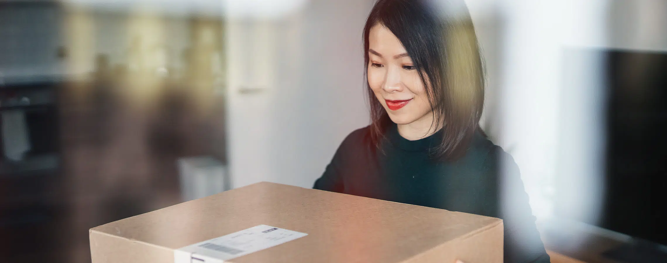 A woman is holding a cardboard box indoors smiling gently in a softly-lit room.