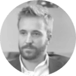 A person with facial hair sits indoors wearing a suit jacket in a grayscale image.