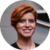 A person with short red hair smiles in an indoor setting with a gray wall and partial logo behind.