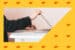 A hand opens a laptop on a white table framed by a bright yellow patterned border.