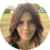 A person with wavy hair is outdoors standing on grass with trees and buildings in the background.