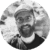 A bearded person smiles wearing glasses and a cap with a blurred outdoor background.