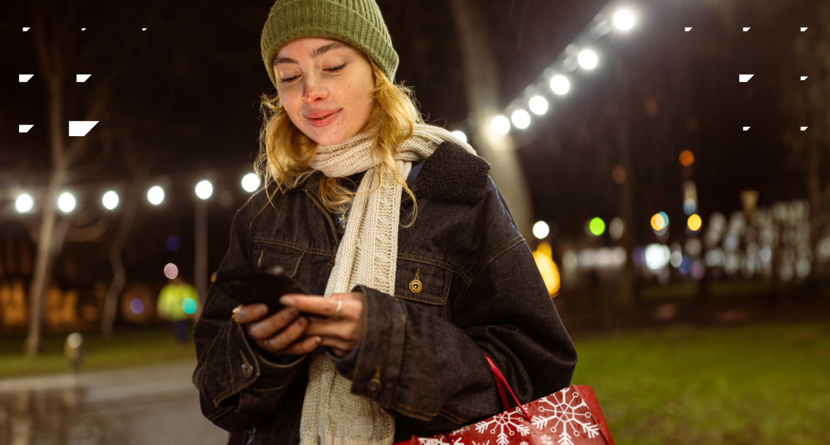 Young Woman walking outside in winter, holding a phone surrounded by holiday lights