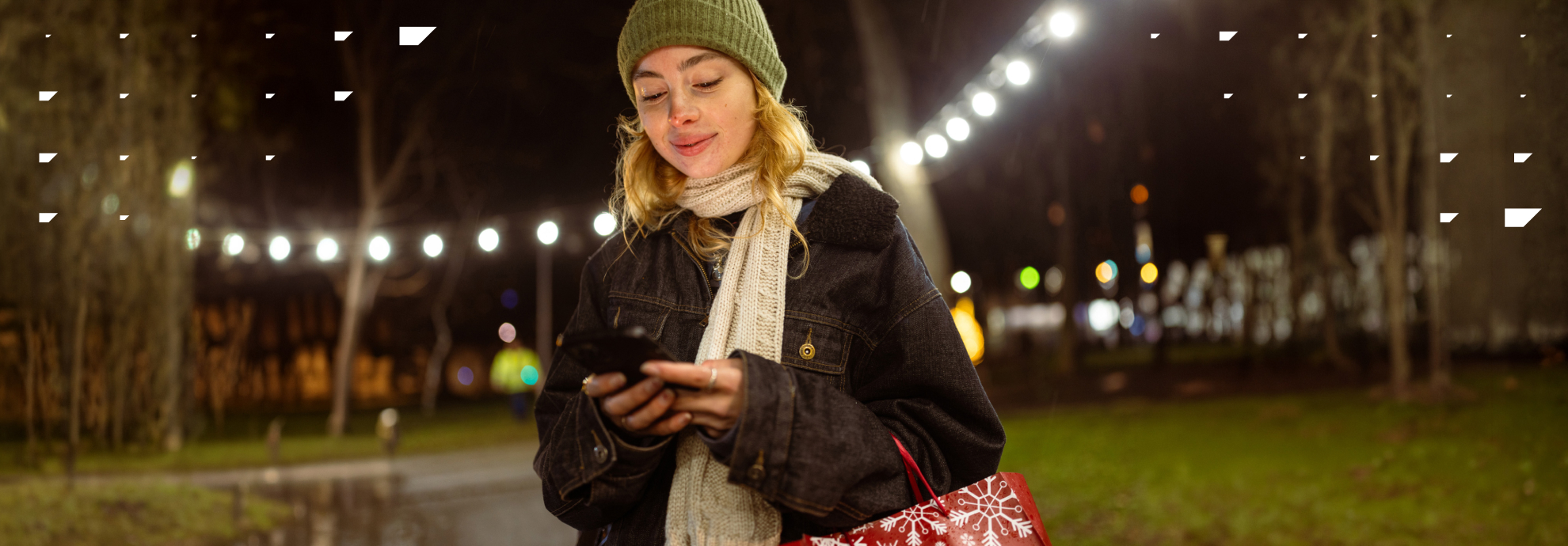 Young Woman walking outside in winter, holding a phone surrounded by holiday lights